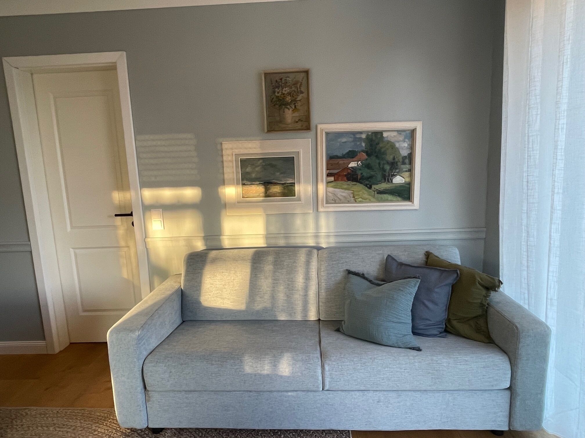 A living room with a light grey sofa, two framed artworks on the wall, and a door to the left.