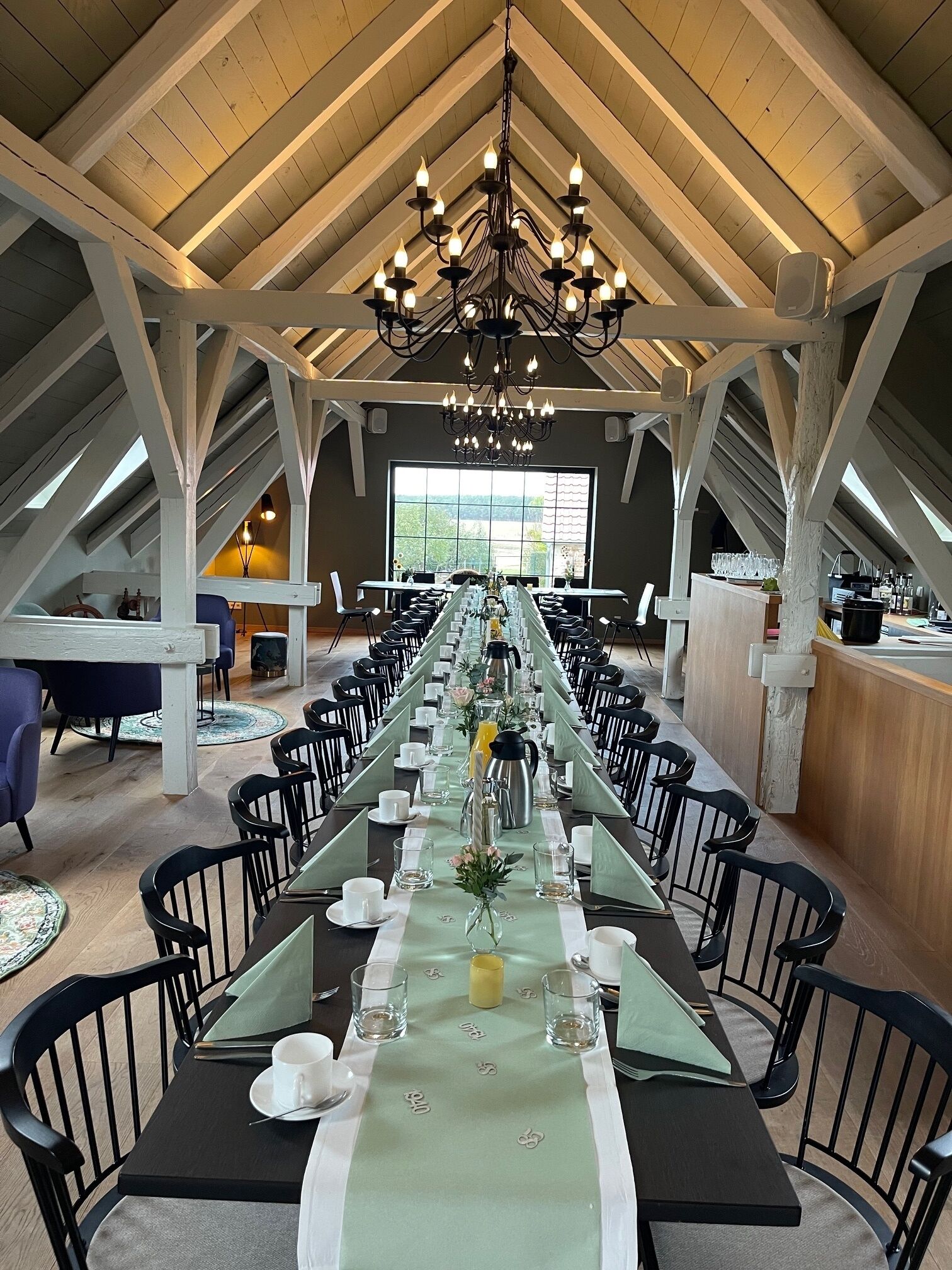 A long dining table set for a meal in a high-ceilinged room with wooden beams and a chandelier.