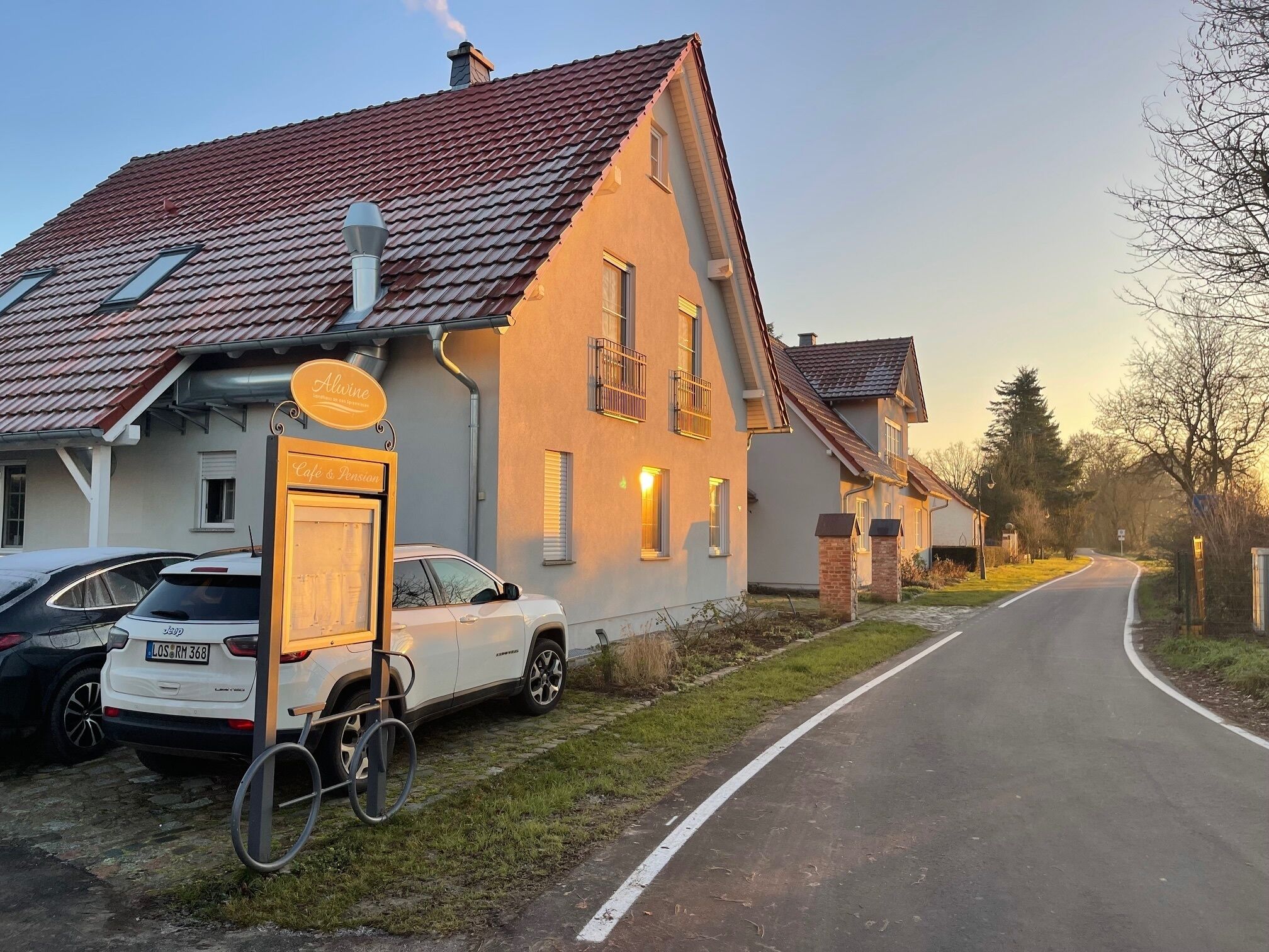 A two-story house with a red-tiled roof, a signboard for 'Alvine Cafe & Pension', and a parked white car with a trailer.