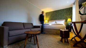 Una camera d'albergo con un divano grigio, un tavolino rotondo, un letto con due cuscini, un comodino con una lampada e una carta da parati decorata.