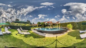 Una piscina circular con sillones de descanso y sombrillas, rodeada por un césped cuidado y setos.