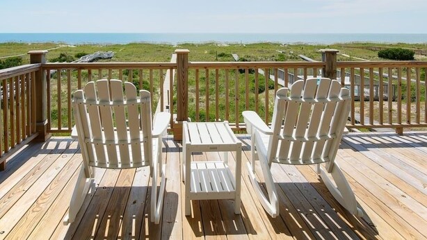 A wooden deck with two Adirondack chairs facing a beach view.