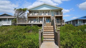 A two-story beachfront house with a wooden staircase leading to a balcony, surrounded by lush greenery.