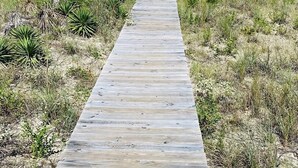 A wooden boardwalk leading to beachfront houses with balconies, surrounded by dry grass and shrubs.