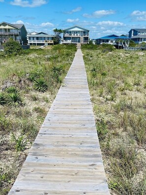 En träpromenad som leder till hus vid stranden, husen har balkonger och omges av torrt gräs och buskar.