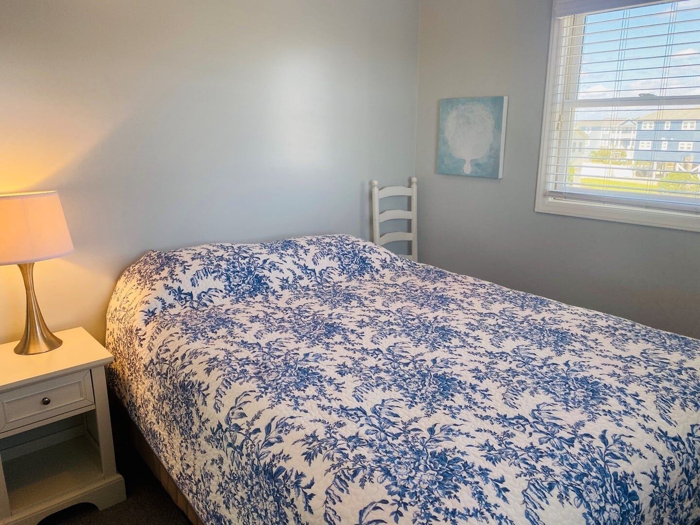 A bedroom with a bed, a nightstand, a lamp, a chair, and a window with blinds.