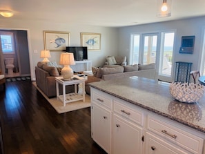 A modern living room with a sofa, a coffee table, and a television. There is a kitchen area with a countertop and cabinets.