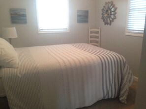 A bedroom with a bed, a chair, two windows, and wall art. - Dune Deck South -  4 Bedroom Home by RedAwning (Supply)