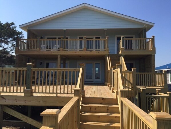 A two-story building with a balcony on the upper floor, a wooden deck with stairs, and a clear sky. - Dune Deck South -  4 Bedroom Home by RedAwning (Supply)