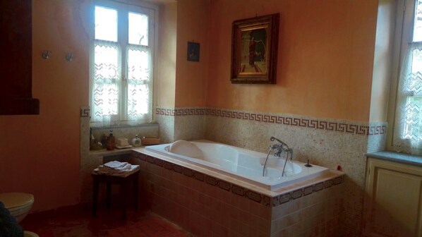 A bathroom with a bathtub, a window with lace curtains, a small table with towels, and a framed picture on the wall. - Villa Mirano B&B (Piossasco)