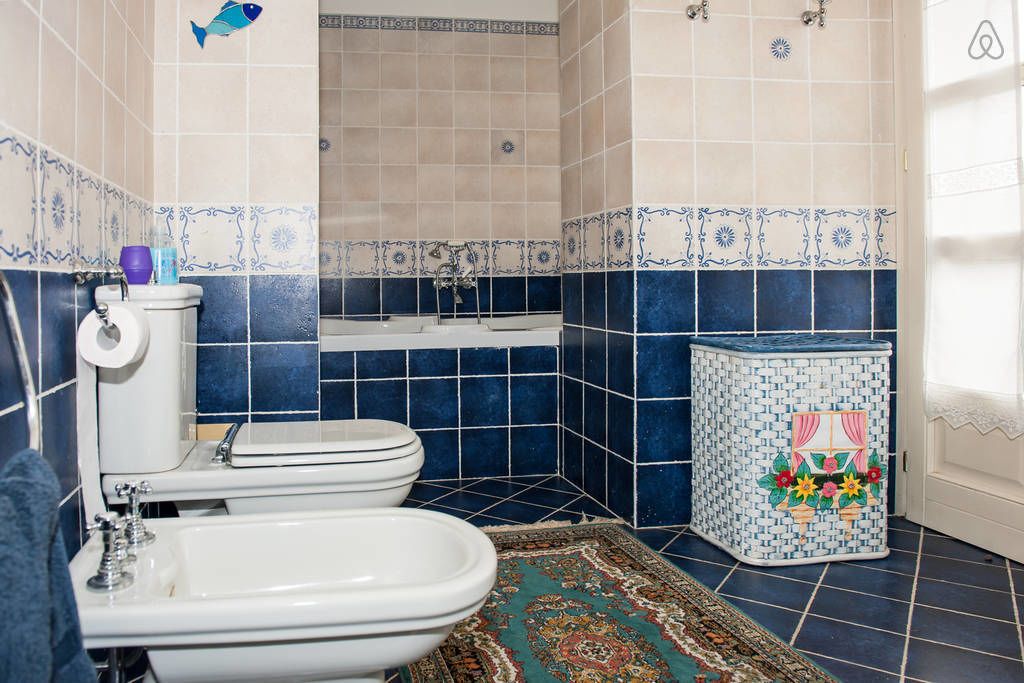 A bathroom with a toilet, bidet, and sink, a decorative rug, and a small cabinet with a floral design.