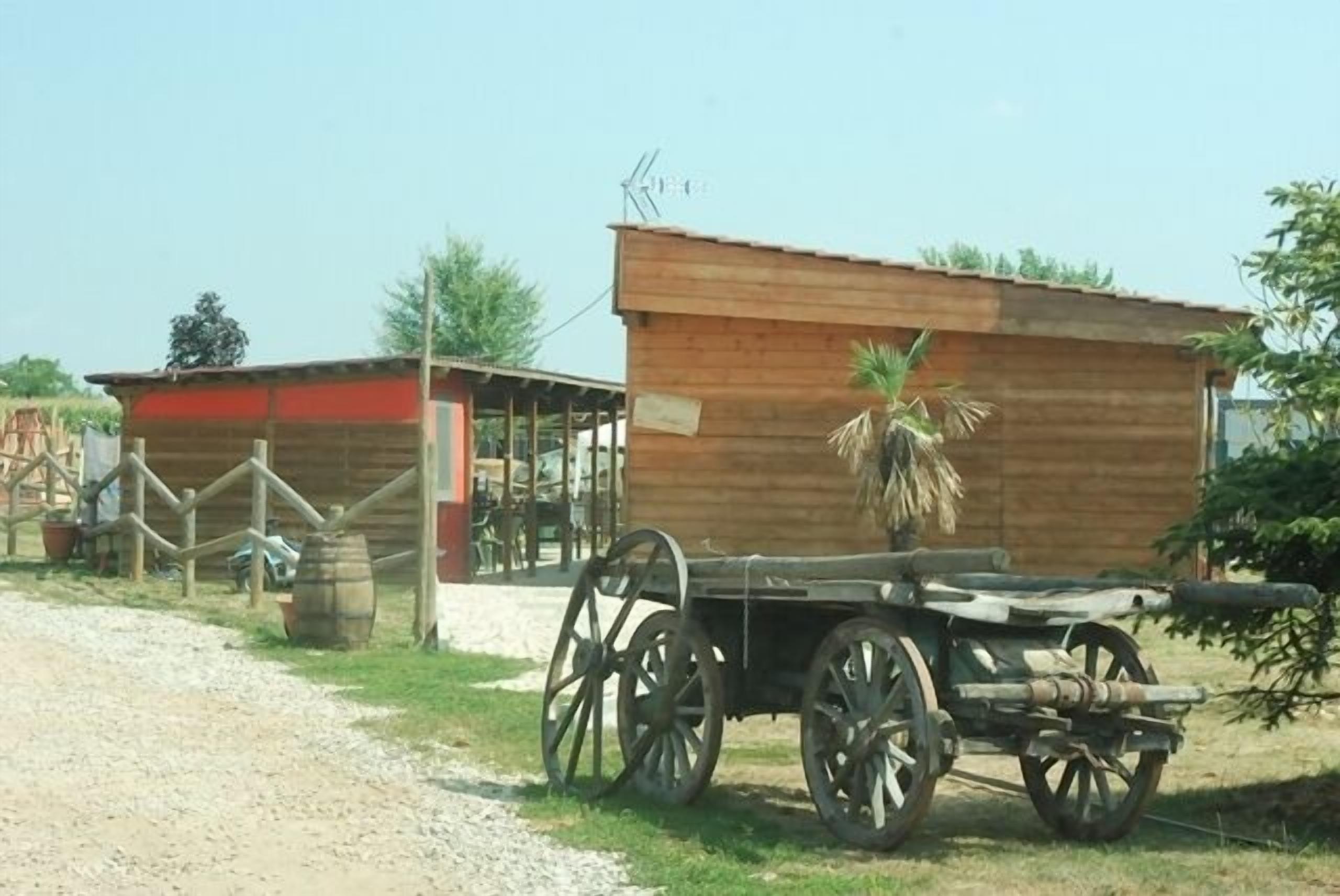 Un edificio rústico de madera con techo de paja, un carruaje de madera con ruedas grandes y un pequeño corralado con una puerta.