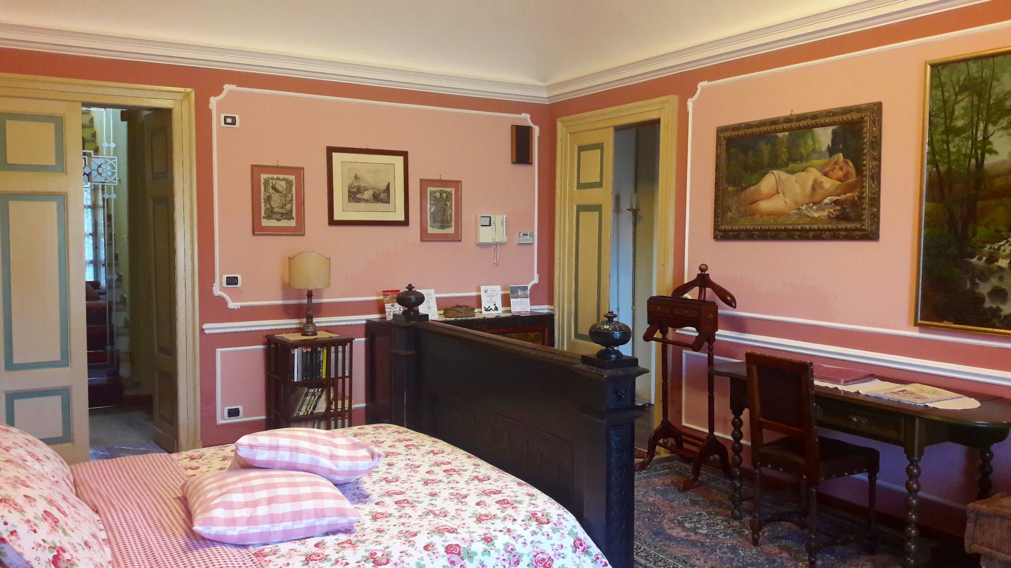 A room with a bed, a desk, and a chair. There are framed pictures on the walls and a painting above the desk.