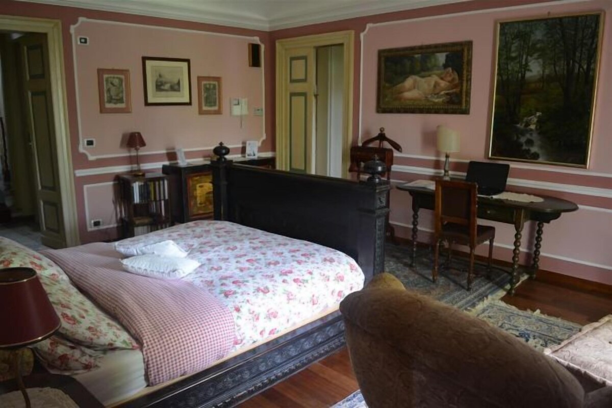 A bedroom with a bed, a desk, and a chair. There are framed pictures on the wall and a painting above the desk.