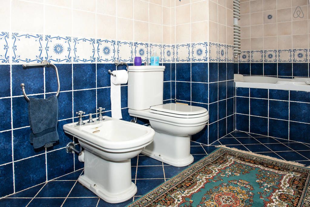 A bathroom with blue tiles, a bidet, a toilet, a towel rack, and a small shelf.