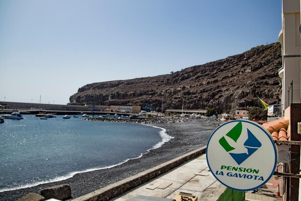 Escena costera con playa, acantilados rocosos y un puerto deportivo con barcos. Se observa un letrero que dice 'Pension La Gaviota'.