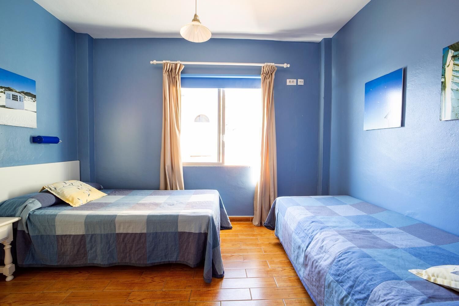 A bedroom with two beds, a window with curtains, a bedside table, and a wall-mounted light fixture.