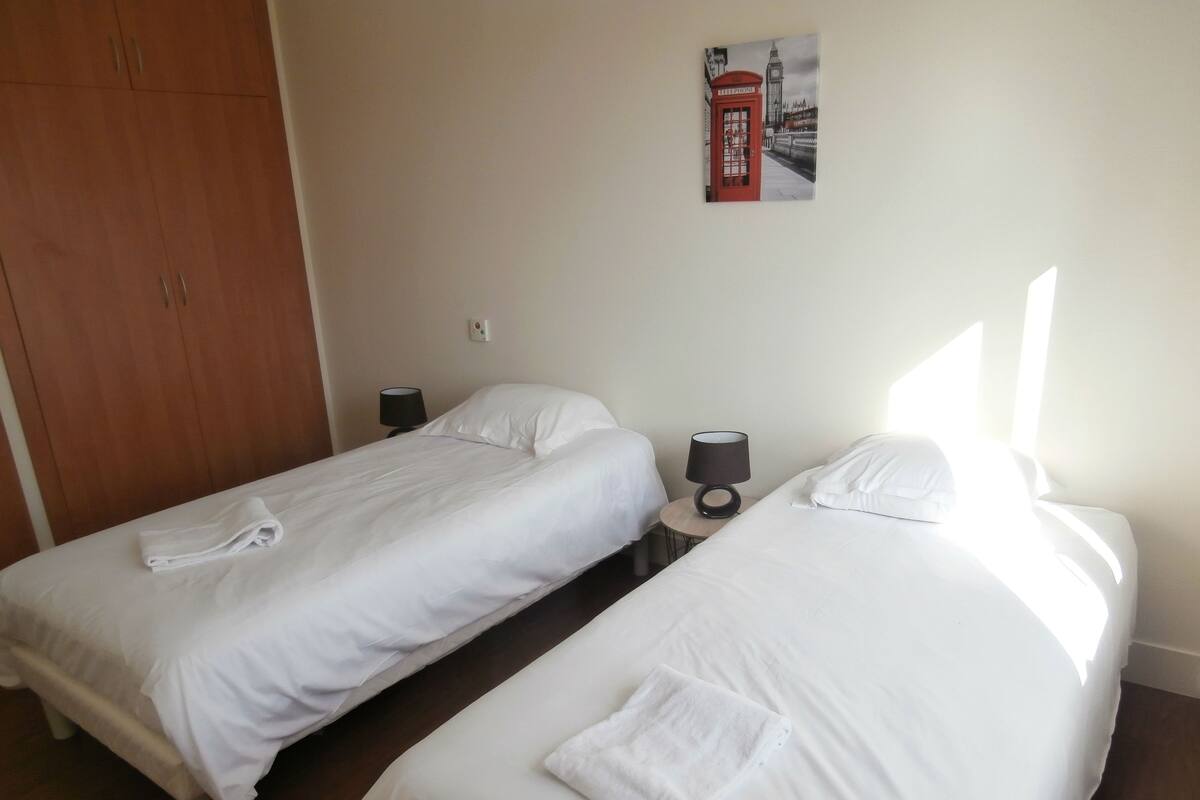 A hotel room with two single beds, a wooden wardrobe, a small round table with a lamp, and a poster on the wall.