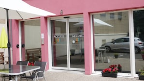 A pink building with glass doors labeled "ACCUEIL" and "OBEO RESIDENCES." There is a table and chairs outside, and a car is parked in the background.
