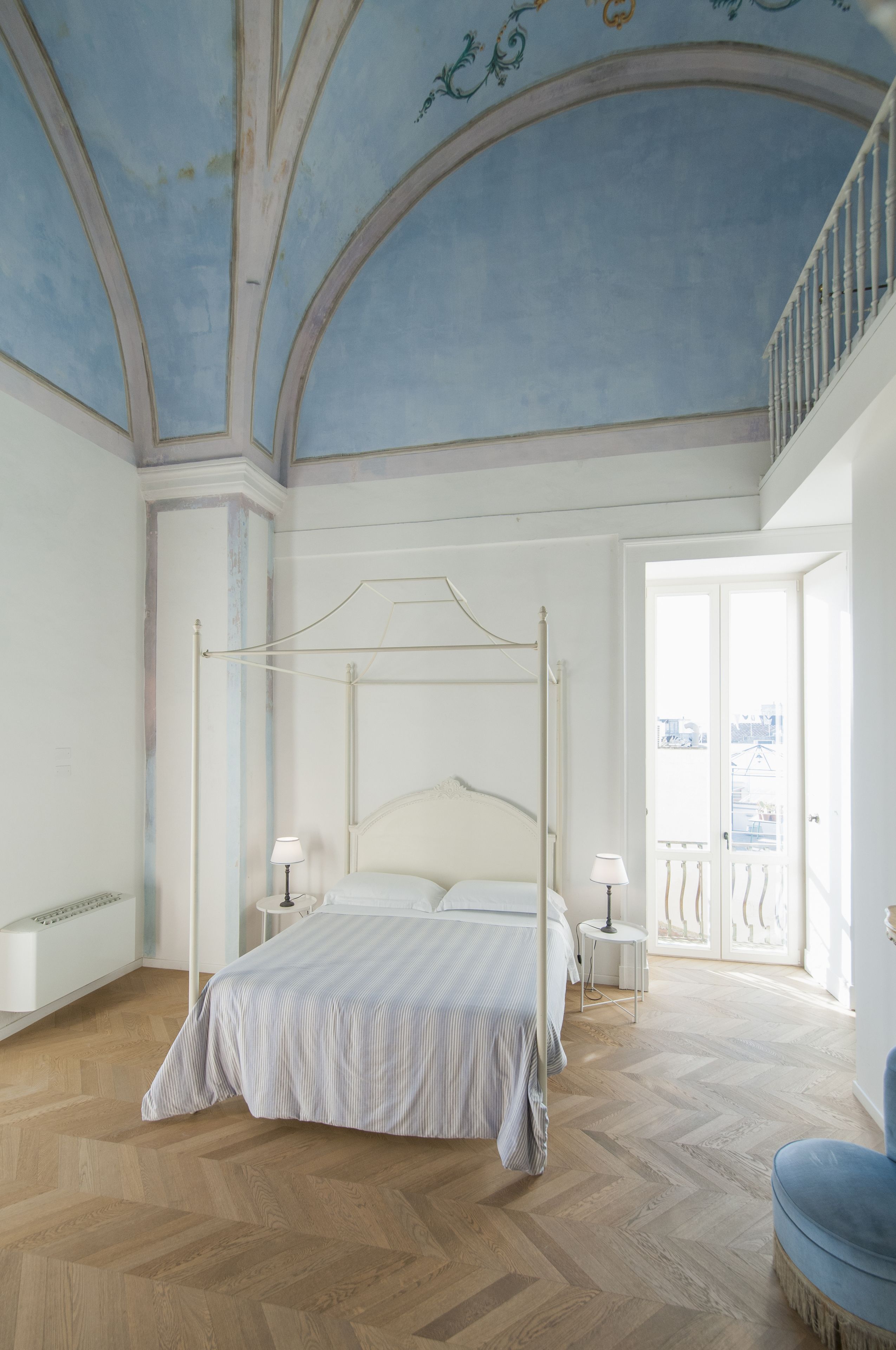 A spacious bedroom with a four-poster bed, a balcony with a railing, and a view of the cityscape.