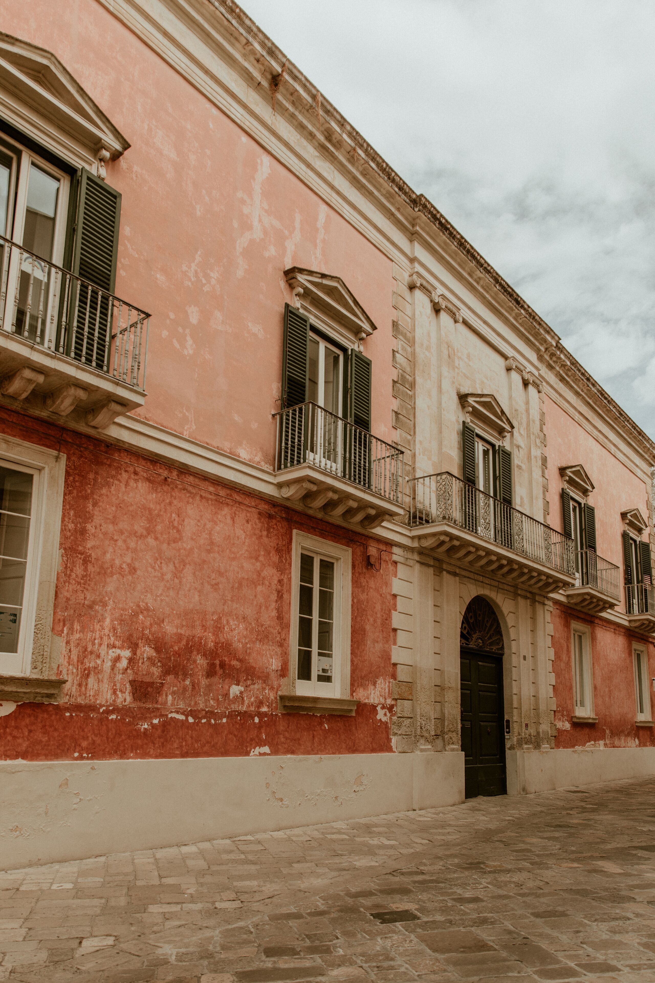 A pink building with a central entrance, multiple windows with shutters, and balconies.