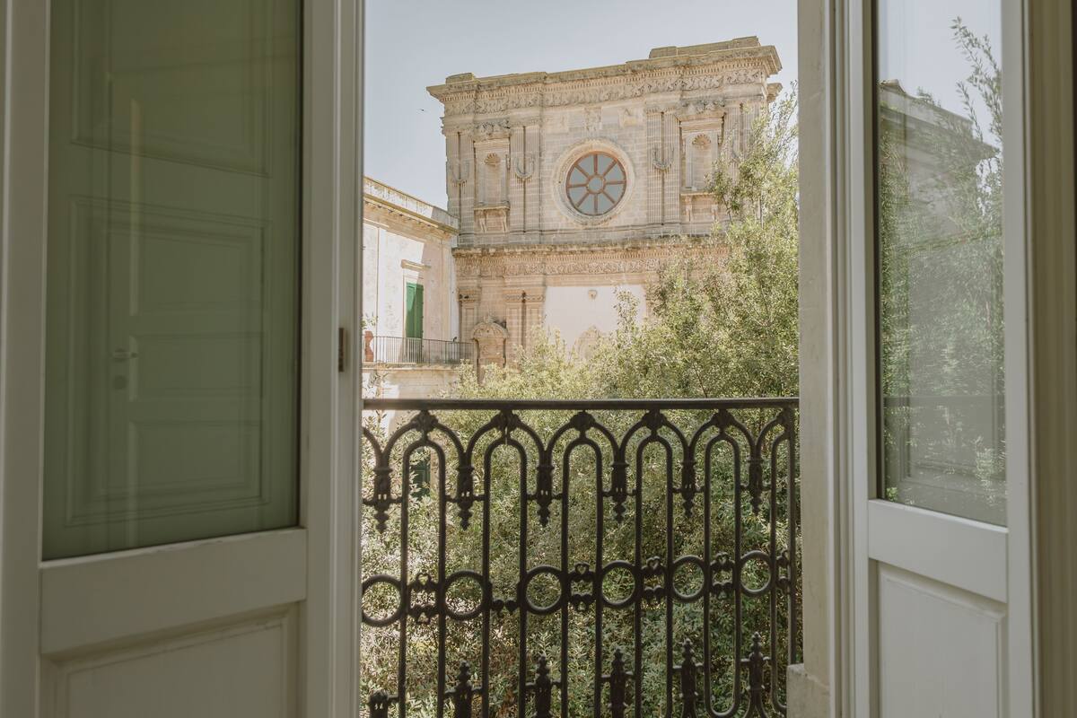 Vista attraverso una finestra con un balcone, che mostra un edificio storico dall'architettura elaborata e una grande finestra circolare.
