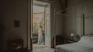A bedroom with a large bed, a nightstand, a chair, and an open door leading to a balcony. - Relais Monastero Santa Teresa (Nardò)