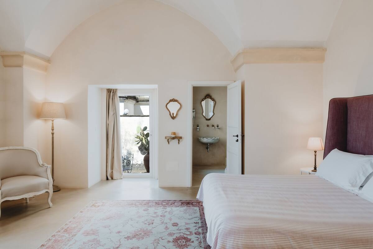 Una camera da letto con un letto, una sedia, una lampada, uno specchio e una porta che dà su un bagno.