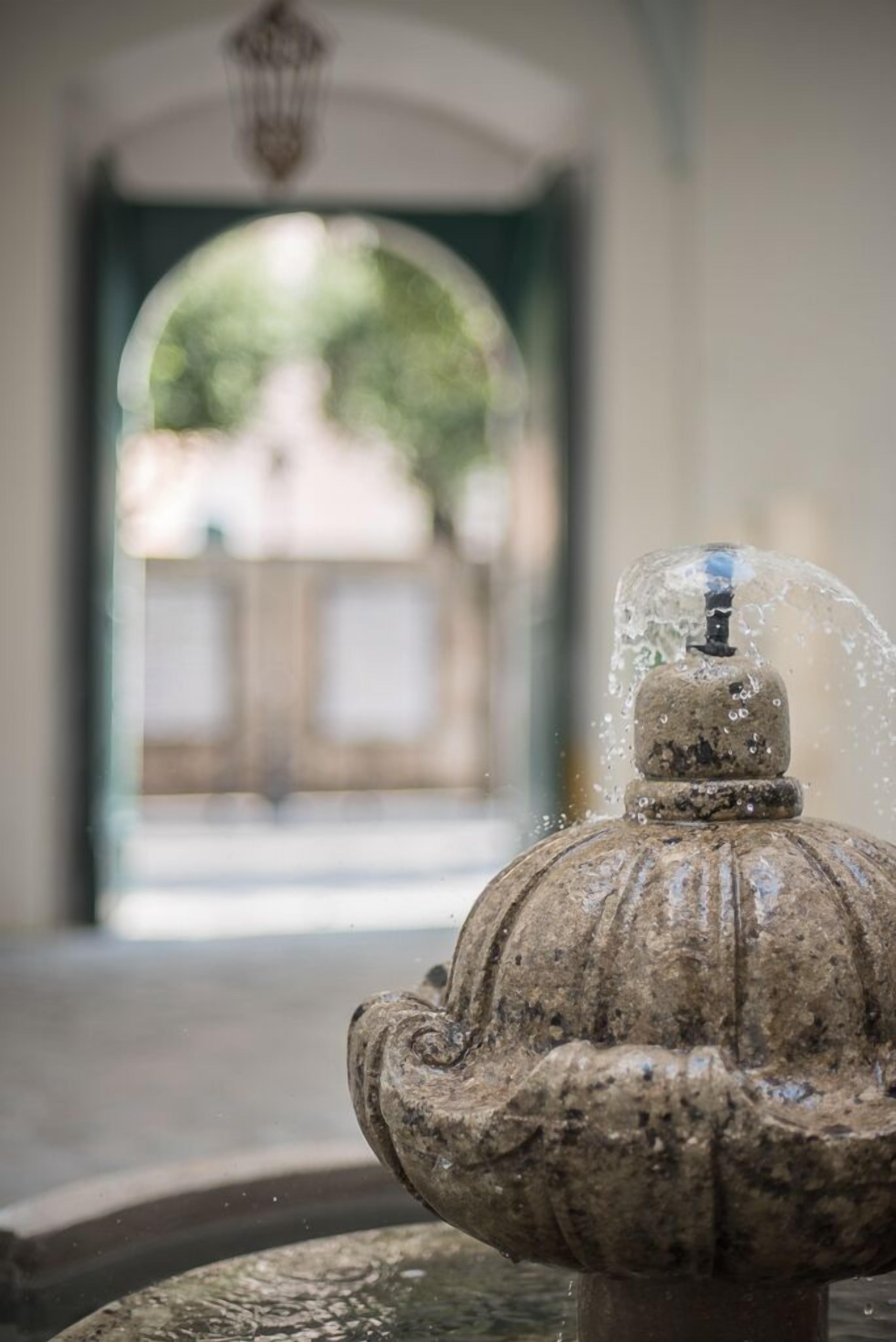 A stone fountain with water spouting from its top, set in a courtyard with an arched entrance in the background.