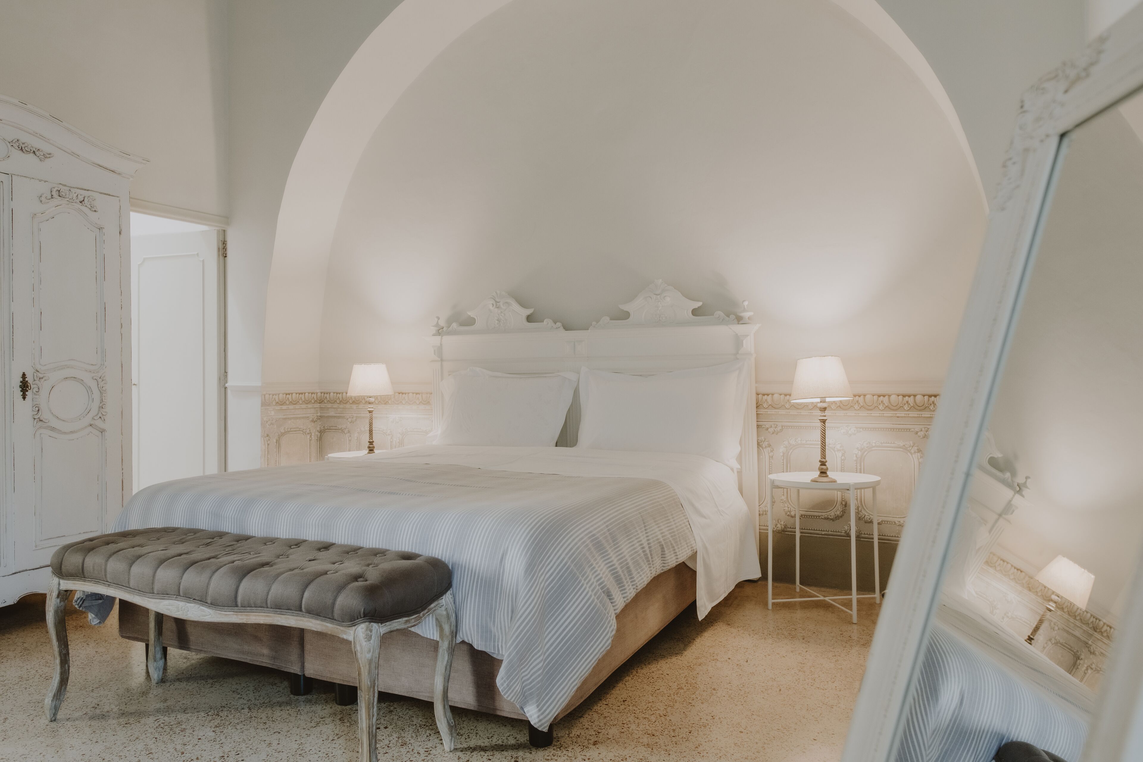 A bedroom with a large bed, a bench, a small table, and a mirror.