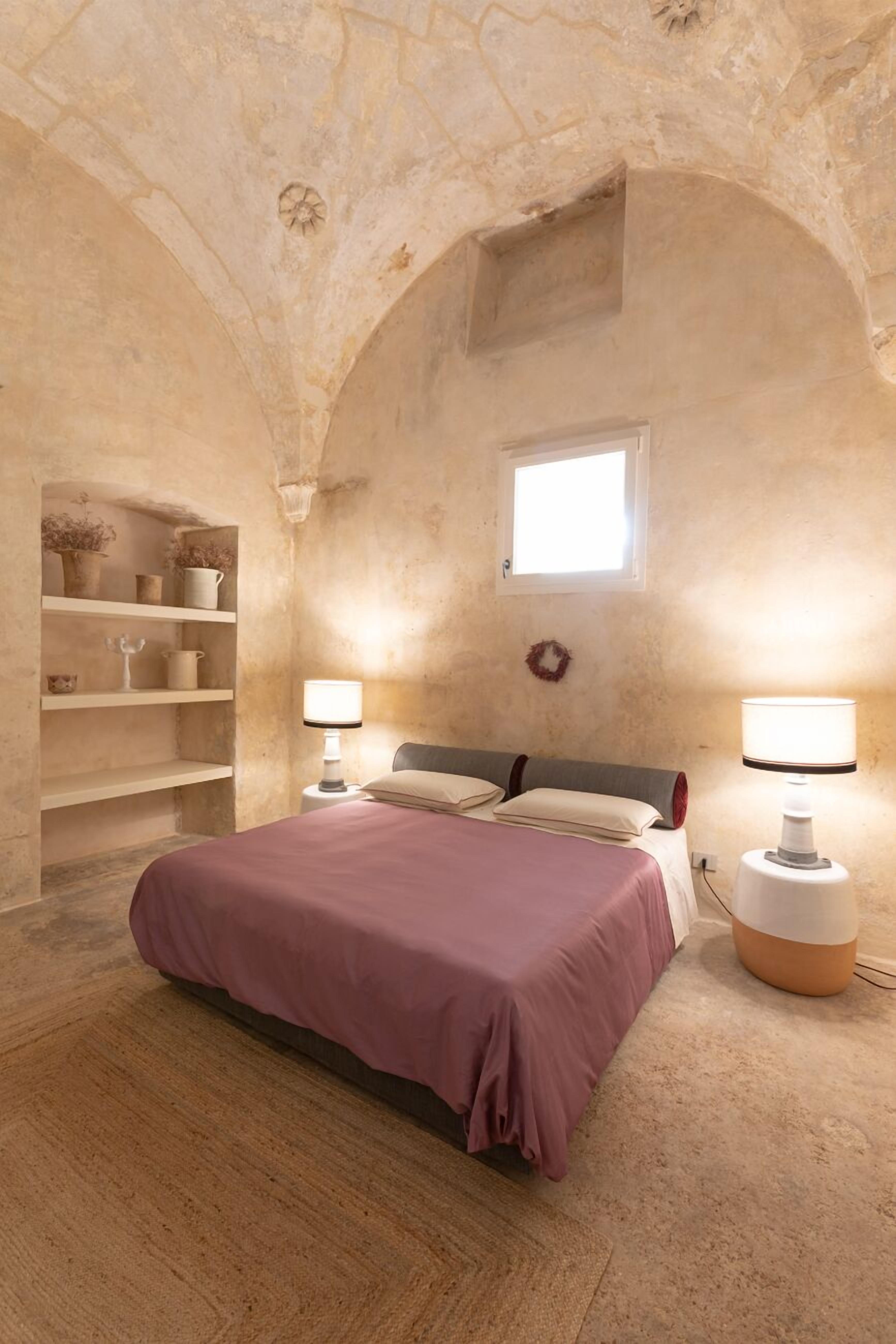Un dormitorio con cama, dos lámparas de noche, una ventana y una estantería con objetos decorativos.