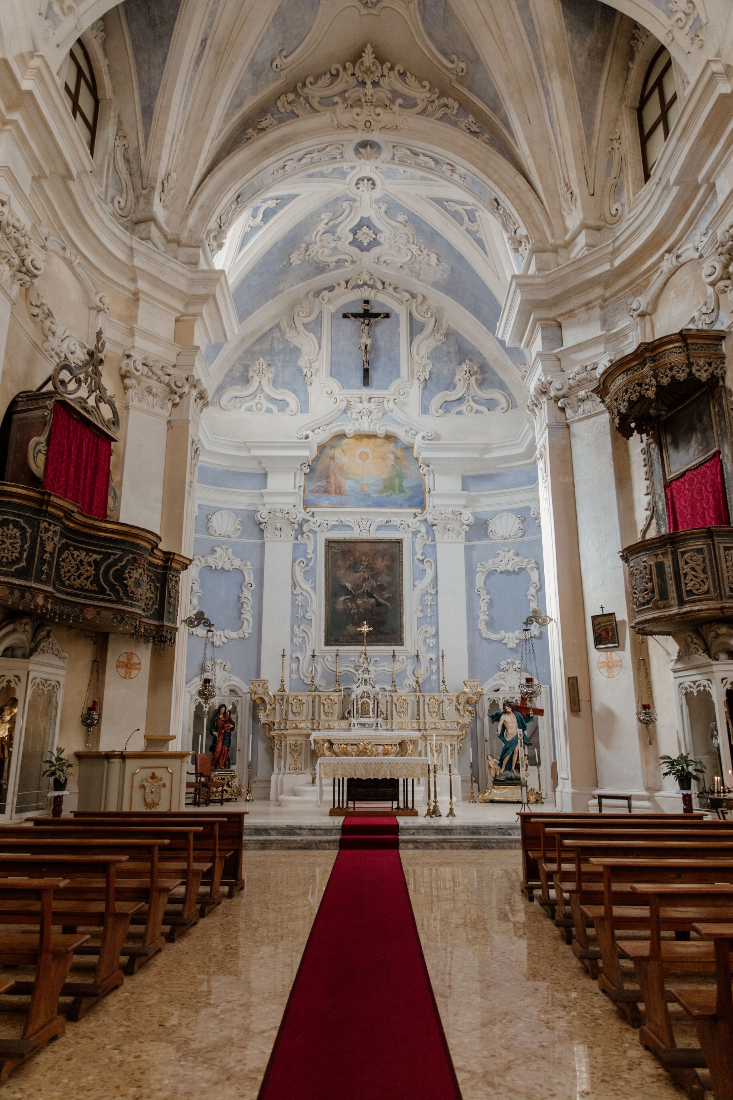 An ornate church interior with a red carpet leading to the altar, wooden pews, and elaborate ceiling decorations.