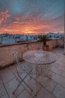 Terraza en la azotea con una mesa y sillas de metal, una planta en maceta y vistas a la ciudad al atardecer.