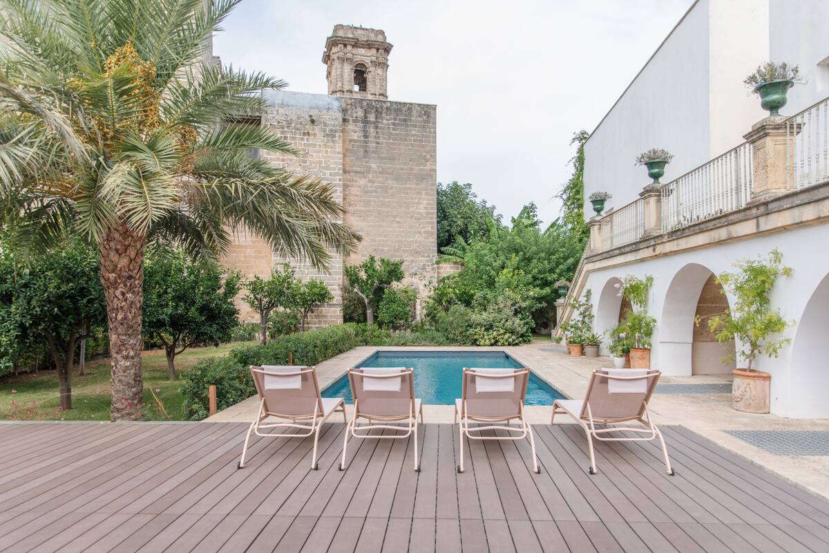Area piscina con lettini, una palma e un edificio storico sullo sfondo.
