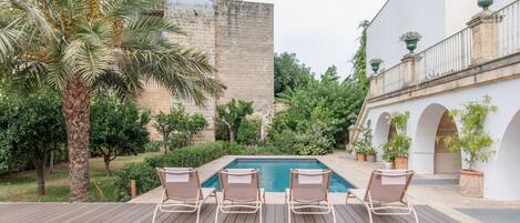 Zona de piscina con sillones de descanso, una palmera y un edificio histórico al fondo.
