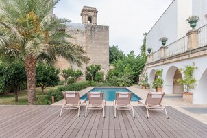 Zona de piscina con sillones de descanso, una palmera y un edificio histórico al fondo.