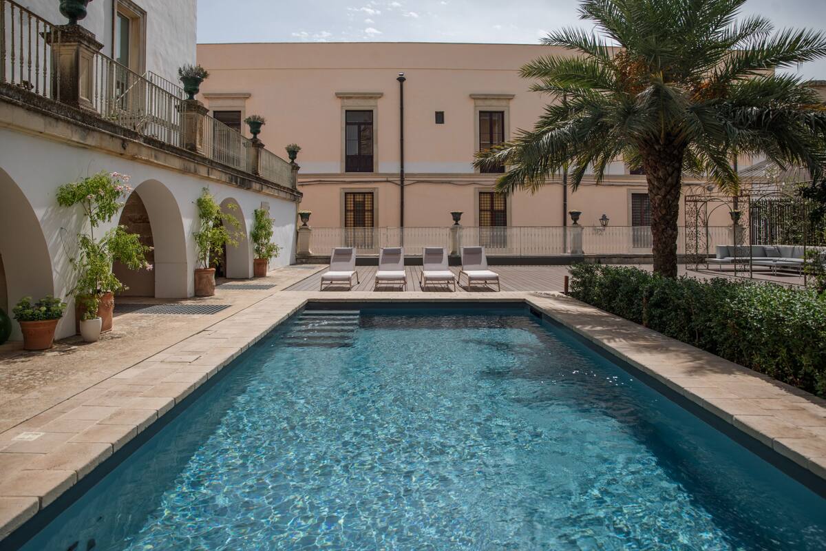 Una piscina rettangolare con acqua cristallina, circondata da un solarium piastrellato e piante in vaso.