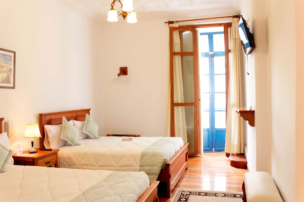 A hotel room with two beds, a wooden floor, a TV mounted on the wall, and a door leading outside.