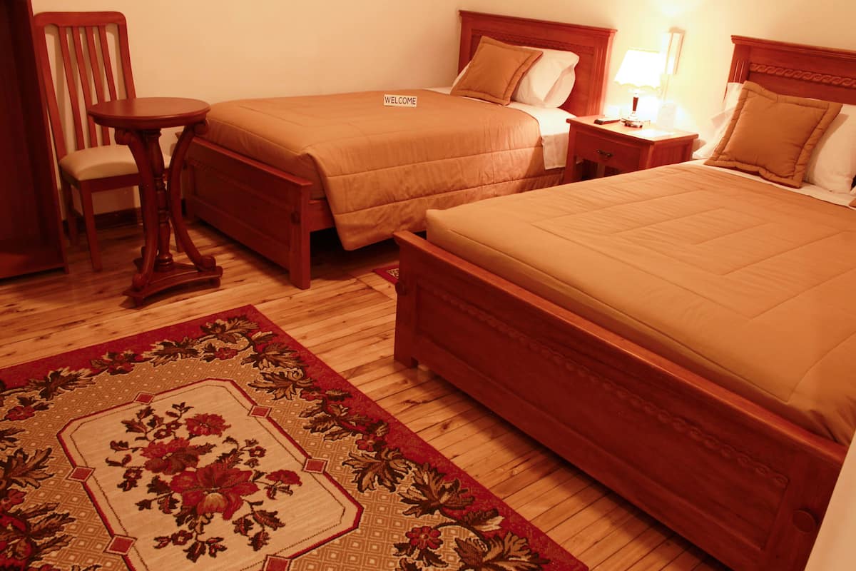 A hotel room with two single beds, a wooden chair, a small round table, and a nightstand with a lamp.