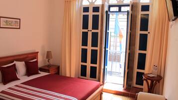 A hotel room with a large bed, a bedside table with a lamp, a small table, and a chair. There is a door leading to a balcony.