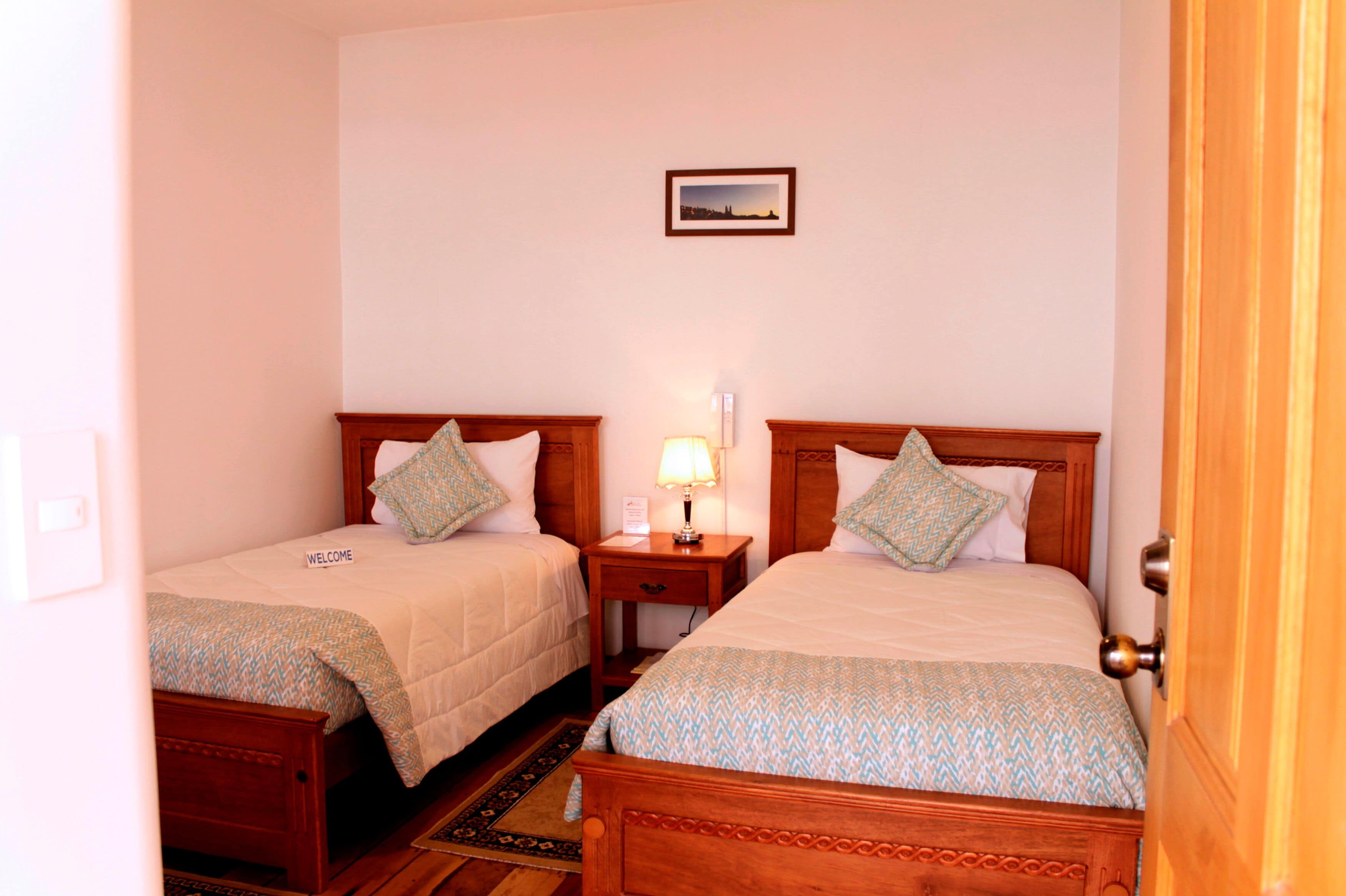 Habitación de hotel con dos camas, una mesita de noche con lámpara, un cuadro en la pared y una puerta de madera.