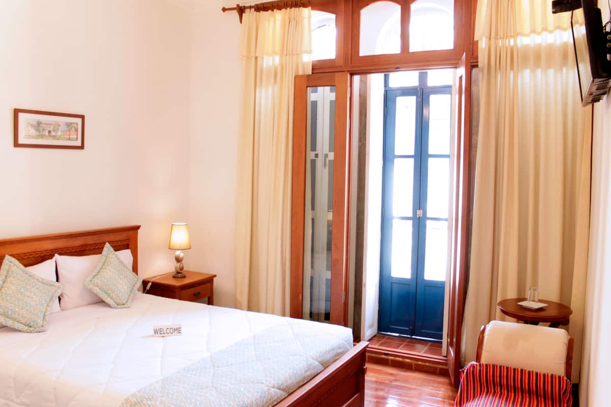 A hotel room with a bed, a nightstand with a lamp, a window with curtains, and a door leading outside.