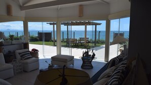 A spacious living room with a large window overlooking the sea, a patio area with a pergola, and a view of the ocean. - Casa do Mar Buzios (Búzios)