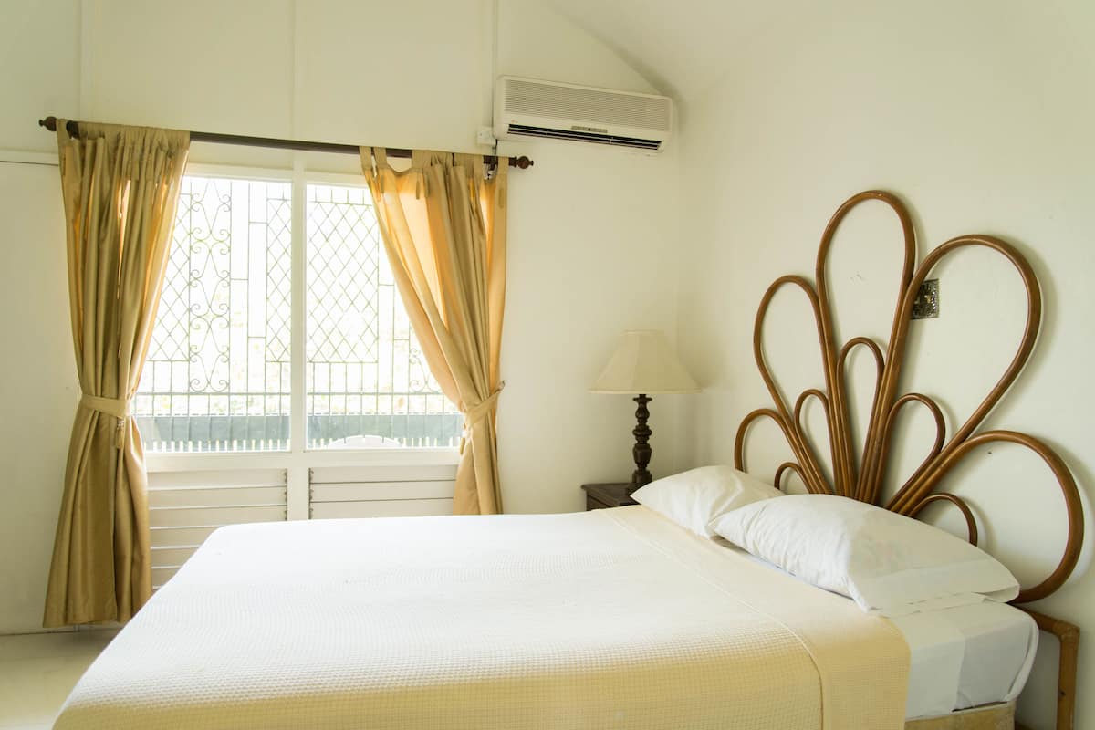 A bedroom with a bed, curtains, an air conditioning unit, a nightstand with a lamp, and a decorative headboard.