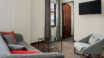 A living room with a grey sofa, a grey armchair, a glass coffee table, and a flat-screen TV mounted on the wall.