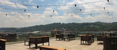 Terraza en la azotea con mobiliario exterior, luces colgantes y vistas a un paisaje urbano y colinas.