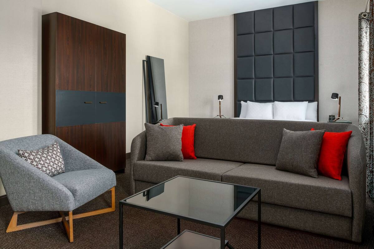 Ein modernes Hotelzimmer mit grauem Sofa, grauem Sessel, einem Glastisch und einem dunklen Kopfteil sowie einem großen Spiegel.