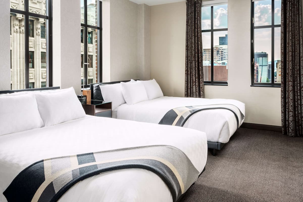 Ein Hotelzimmer mit zwei Betten, großen Fenstern, Teppichboden und Blick auf die Stadt.