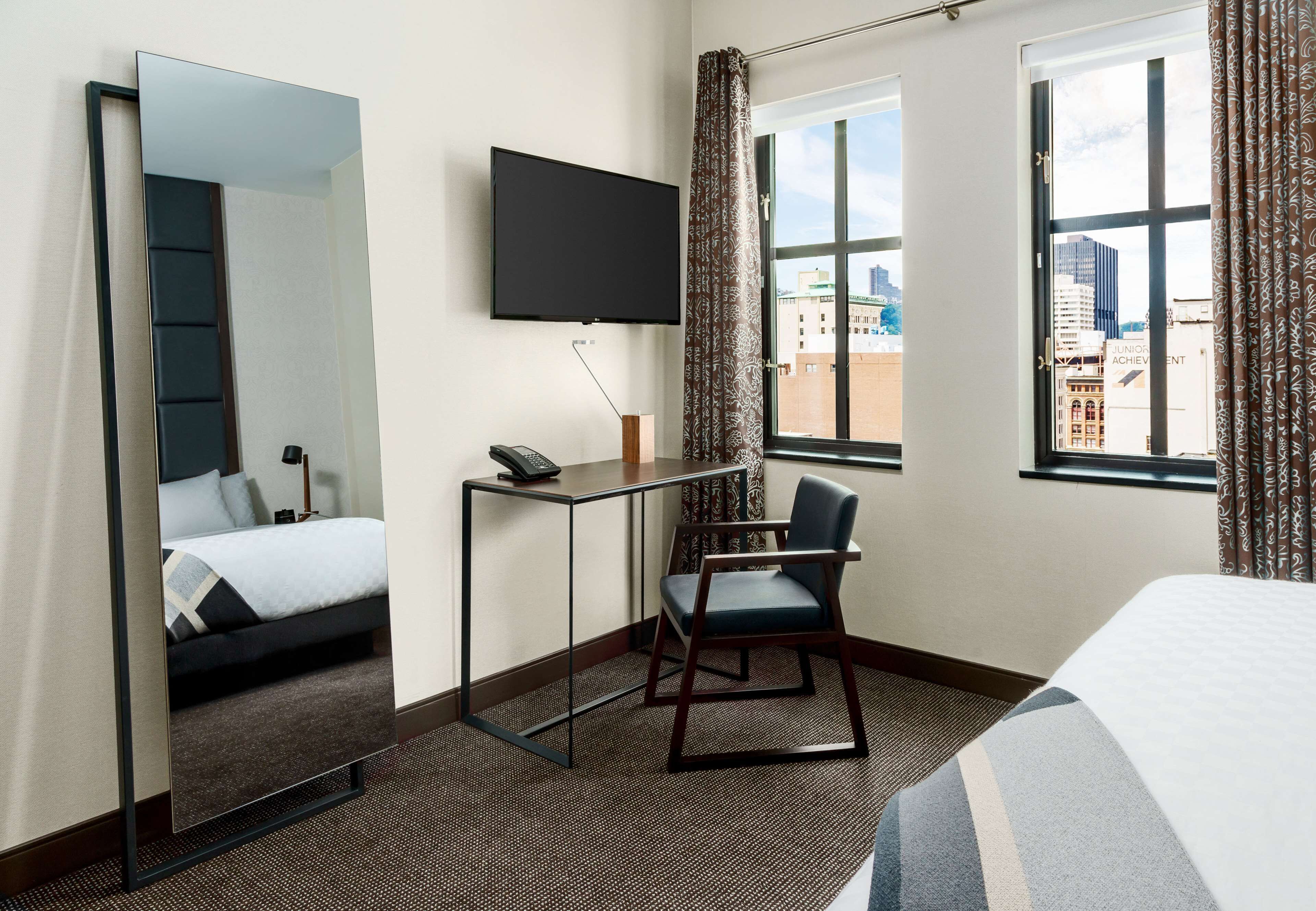 A hotel room with a bed, a desk with a chair, a TV mounted on the wall, and a window with a city view.