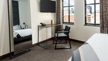Habitación de hotel con cama, escritorio con silla, televisor montado en la pared y ventana con vista a la ciudad.